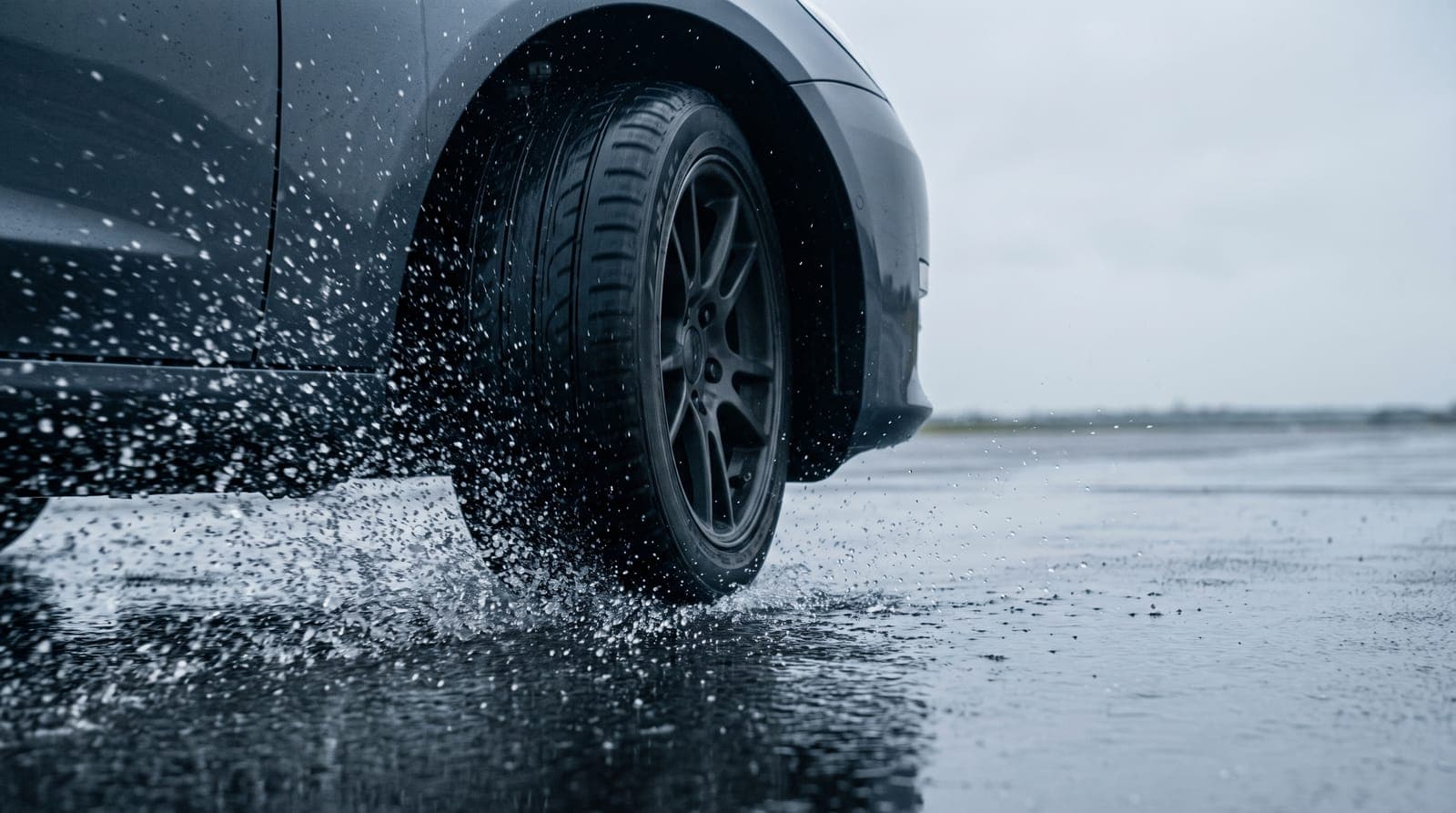 Rueda delantera de un coche frenando sobre asfalto encharcado, con el agua saliendo despedida de la huella del neumático